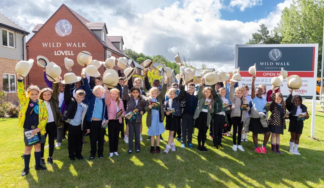 A group of children in a grassy area, many holding up safari hats, with new houses and a 'Wild Walk Lovell' sign in the background.