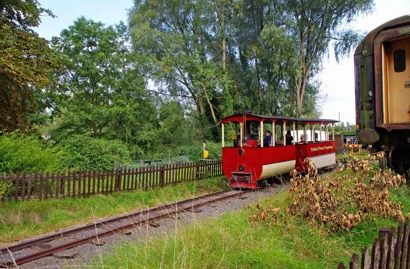 A red and white open-top train, labeled 'Telford Town Tramway', travels along tracks next to a brown picket fence and lush green foliage, with another train car visible on the right.