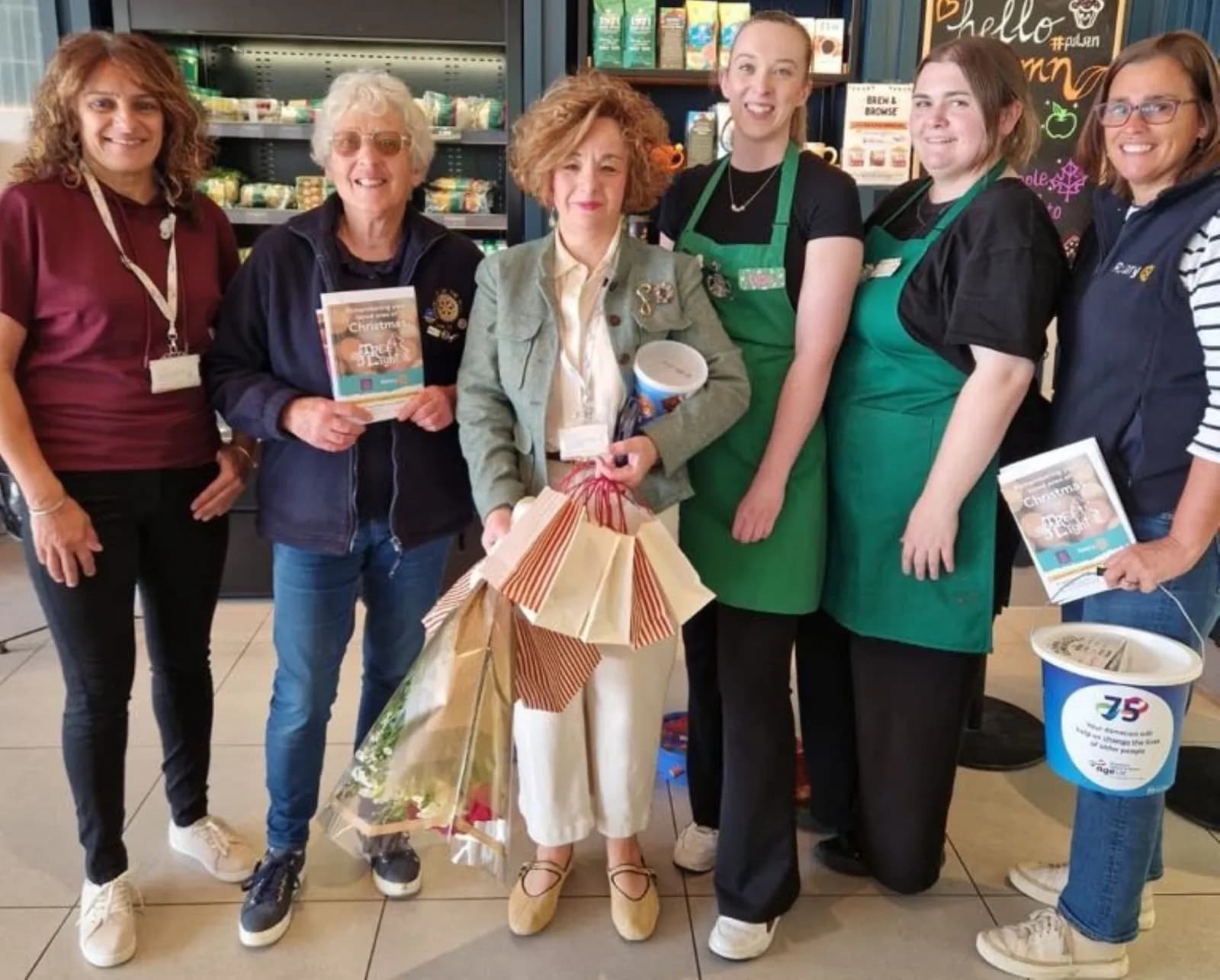 Six women are standing together, three of whom are wearing green aprons. Two women hold books with "Christmas" on the cover, and one woman holds a bouquet of flowers and small gift bags.