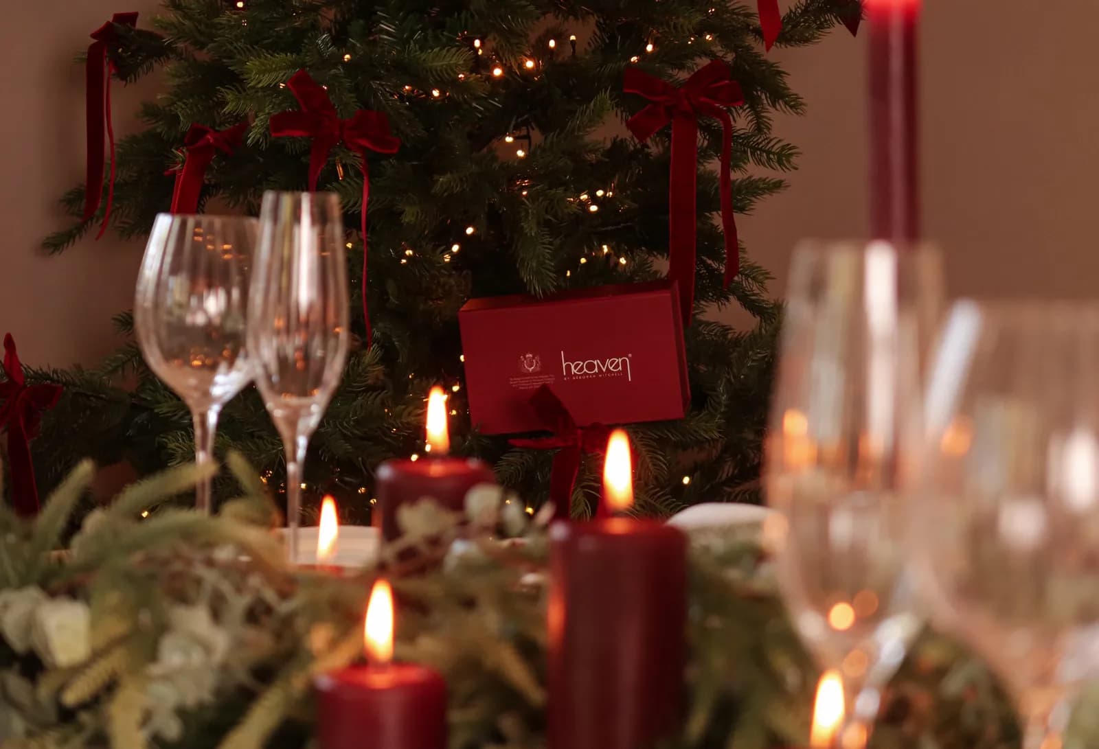 A red box from the brand Heaven hangs from a lit Christmas tree with several wine glasses and lit candles in the foreground.