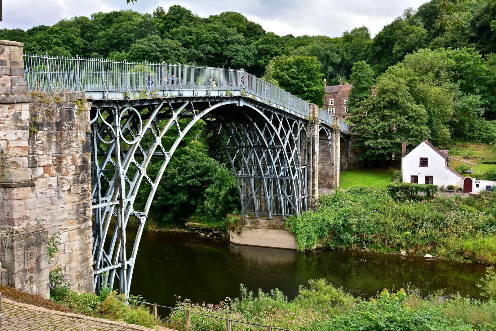 A historic cast-iron bridge with intricate lattice work spans a dark river, connecting lush green banks with distant houses and trees under a cloudy sky.