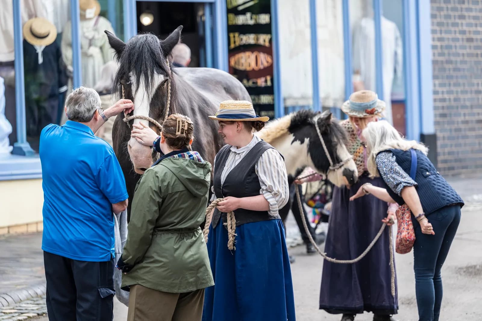 A group of people interact with two horses on a street in front of a shop.