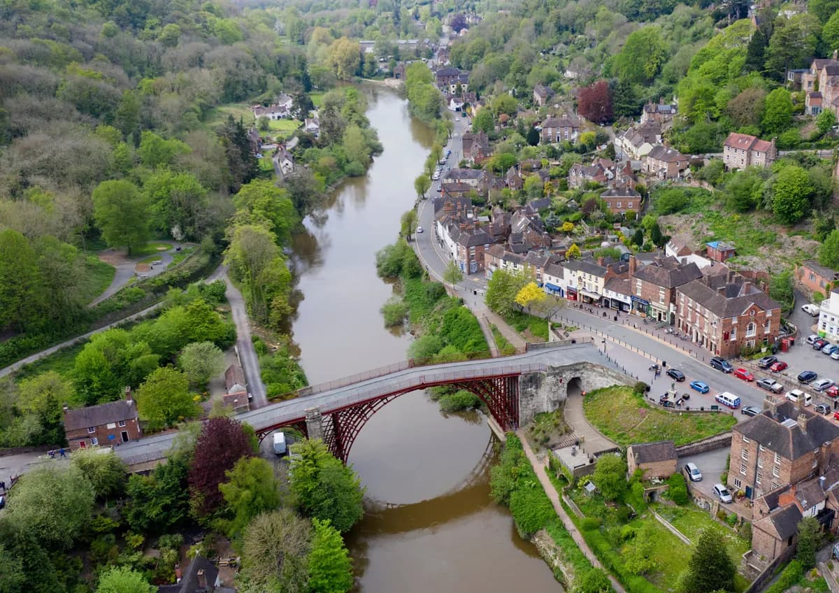 An aerial view shows a river flowing through a forested valley with a town built along one bank and a red metal bridge crossing the river.